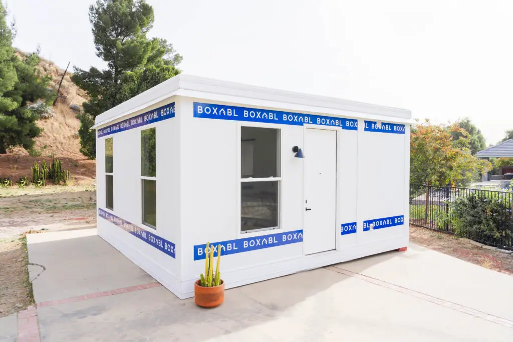 Boxabl modular home showcasing sleek, modern design with large windows and a front door, surrounded by greenery in a San Diego setting, emphasizing efficient and sustainable living solutions.