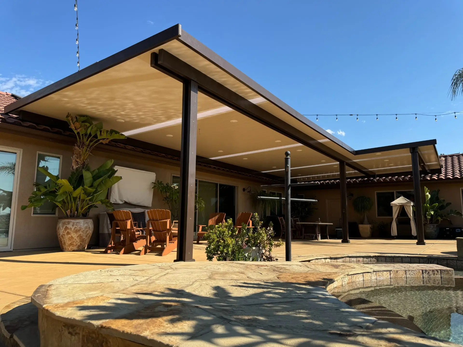 Custom aluminum patio cover installation in residential backyard, featuring modern design, outdoor seating area with wooden chairs, and lush greenery, emphasizing quality craftsmanship and outdoor living space enhancement in Alpine, CA.