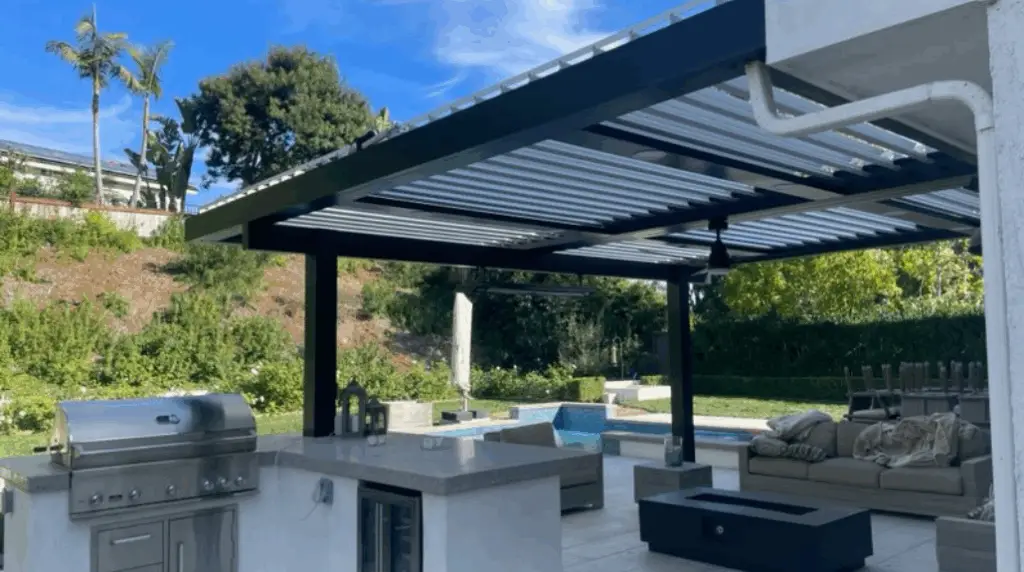 Modern pergola structure with retractable roof, outdoor kitchen featuring stainless steel grill and bar seating, surrounded by lush greenery and a swimming pool, emphasizing professional craftsmanship in outdoor living spaces.